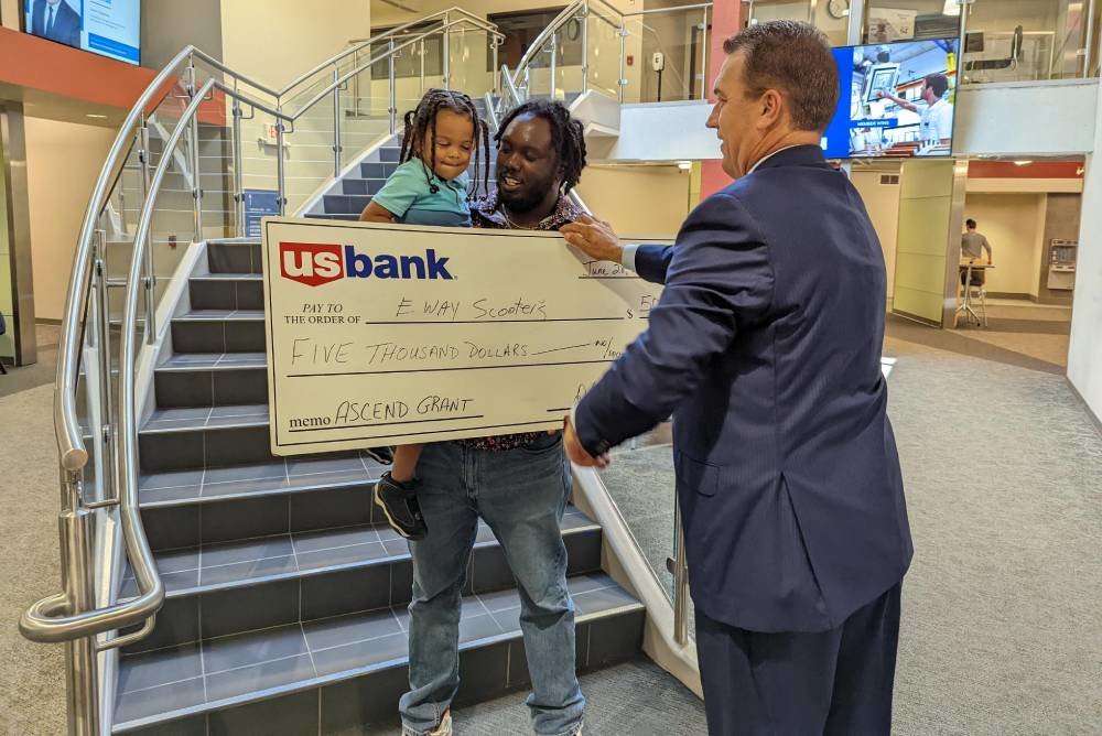 Marquez Williams, owner of Eway Scooters, gets help from 3-year-old son Marquell Williams to heft a check, presented through the Ascend program.